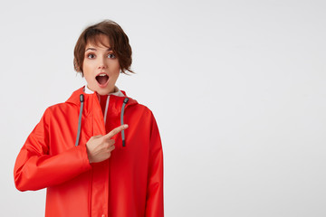 Young nice happy amazed short haired lady in red rain coat, with wide open mouth, hears cool news. Wants to draw your attention, points fingers to the copy space. Standing over white background.