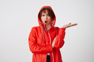 Cute outraged young short haired lady in red rain coat, looks admiringly with wide open mouth in...