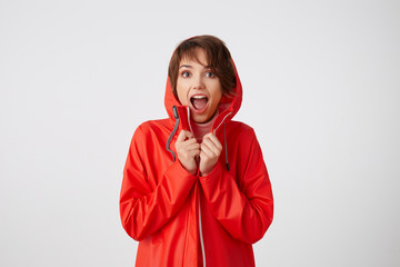 Portrait of young cute surprised short haired woman in red rain coat, looking at the camera with...