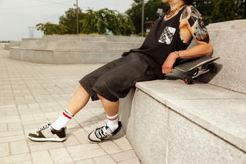 Skateboarder resting after riding at the city's street in cloudly day. Young man in sneakers and...