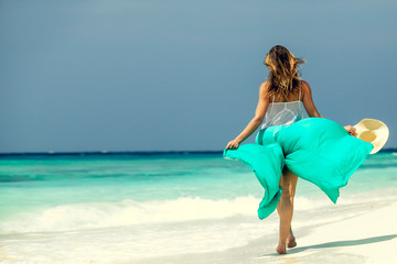 Blonde girl walking in turquoise pareo and straw hat on the beach, ocean in Maldives. Luxury summer beach vacation. Banner panorama landscape. 