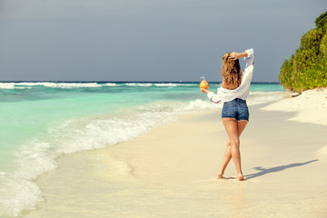 Fototapeta premium Blonde girl in denim shorts with fresh coconut in hand, on beach ocean in Maldives. Summer vacation travel. Banner panorama landscape. 