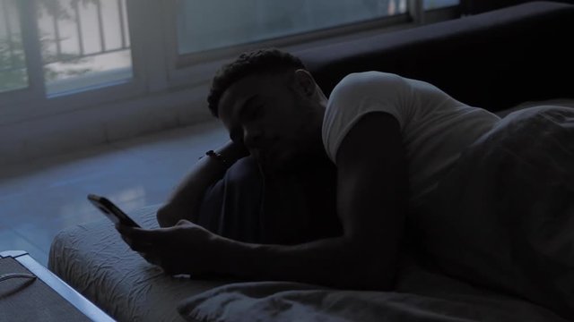 Young afro american man waking up the morning, checking smartphone and getting out of bed. Social network account feed, news, 4K. Communication, social networking, online shopping.