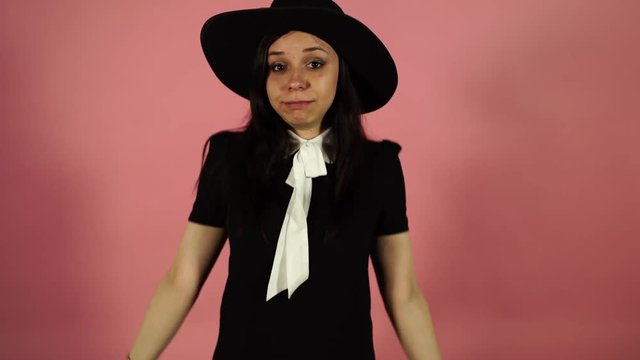 Young female in black dress and hat on a pink background. Attractive woman wearing black outfit standing on pink background in perplexity on a pink background. The confused woman shrugs