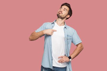 This is me. Portrait of proud haughty handsome bearded young man in blue casual style shirt standing, looking away and pointing himself. indoor studio shot, isolated on pink background. - obrazy, fototapety, plakaty