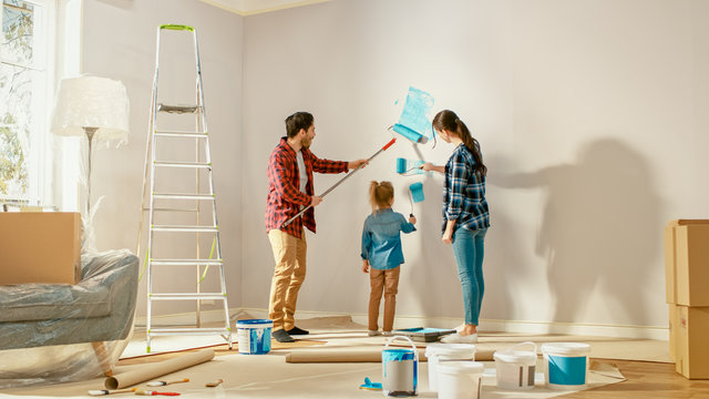 Beautiful Young Family Are Showing How To Paint Walls To Their Adorable Small Daughter. They Paint With Rollers That Are Covered In Light Blue Paint. Room Renovations At Home.