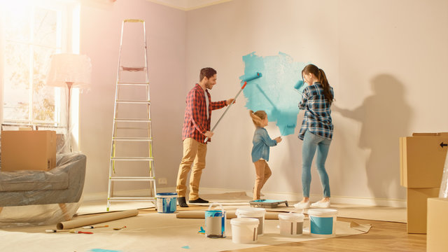 Beautiful Young Family are Showing How to Paint Walls to Their Adorable Small Daughter. They Paint with Rollers that are Covered in Light Blue Paint. Shot with Sun Flare in Renovated Room at Home.