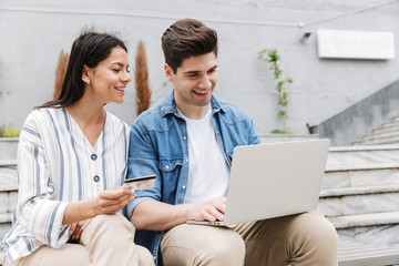 Happy young amazing loving couple business people colleagues outdoors outside using laptop computer holding credit card.