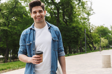 Image of handsome young man drinking takeaway coffee and holding laptop while walking through green...