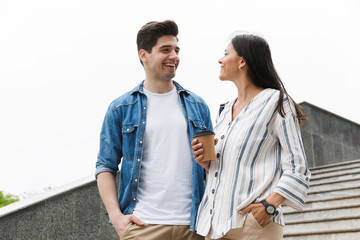 Image of optimistic couple with paper cup smiling and talking while strolling down stairs outdoors