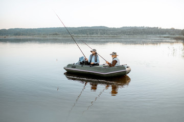 Grandfather with adult son fishing on the inflatable boat on the lake with calm water early in the...