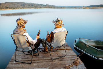 Grandfather with adult son enjoying beer, sitting together on the pier while fishing on the lake early in the morning