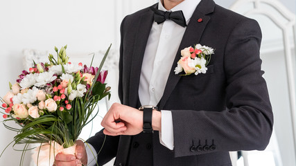 An elegant young man in a black suit and with a clock on his hand and a bouquet of roses.