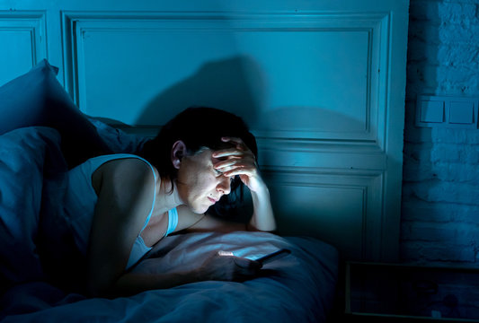 young beautiful woman lying bored in bed using smart phone late at night in a dark bedroom