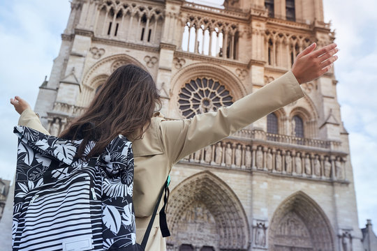 Young happy woman standing in front of the famous Notre Dame cathedral in Paris, hands raised up to the sky
