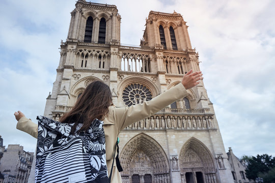 Young happy woman standing in front of the famous Notre Dame cathedral in Paris, hands raised up to the sky