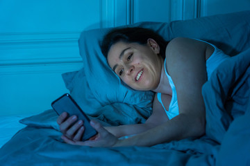 Happy sleepless woman lying in bed on the internet using mobile phone late at night in dark bedroom