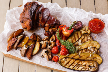Grilled pork knuckle with grilled tomatoes, champignons, vagetable marrow, eggplant, red sweet pepper and baked potatoes and parchment on a wooden cutting board. top view