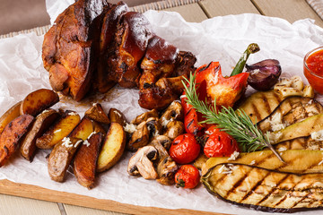 Grilled pork knuckle with grilled tomatoes, champignons, vagetable marrow, eggplant, red sweet pepper and baked potatoes and parchment on a wooden cutting board. top view