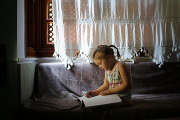 Fototapeta premium Child draws in large notebook on couch by window