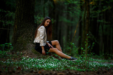 Portrait of a young pretty caucasian teen girl with long hair in casual clothes sitting on the ground in the forest on a summer evening.