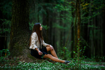 Portrait of a young pretty caucasian teen girl with long hair in casual clothes sitting on the ground in the forest on a summer evening.