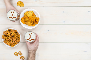 Couple holds glass mug of beer with silhouettes of hockey sticks on beer foam and pretzels and chips on light wooden background. Empty space for text. Top view
