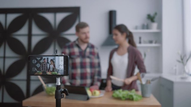 nutritional blog, young vloggers man and woman prepare healthy breakfast with vegetables and greens in kitchen while camera smartphone records video for subscribers at social networks