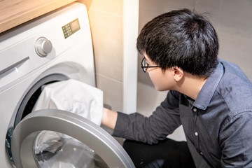 Asian man loading white clothes into front door washing machine in laundry room. Housework or chores concepts