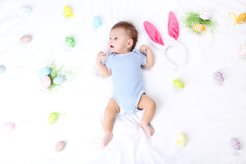 Cute baby with easter eggs and rabbit ears lying on white bed