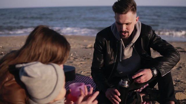 Young stylish family in casualwear sitting on plaid by waterside and having hot drinks at picnic. Mother gives a cup of tea to her son. Cold weather, weekend recreation