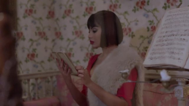 Sexy brunette Arab woman in tight red dress reading a book sitting on a couch, filmed hidden behind a harp in a retro vintage hotel with walls adorned with floral tapestry. Slow motion beauty concept.
