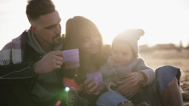 Beautiful scene of young stylish family in casualwear sitting on plaid embracing and enjoying time together, drinking hot drinks from plastic cups. Cold weather, weekend recreation. Lens flares on the