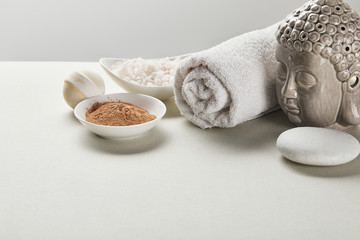 sea salt and clay powder in bowls, cotton towel, stone, bath bomb and Buddha figurine on white table isolated on grey