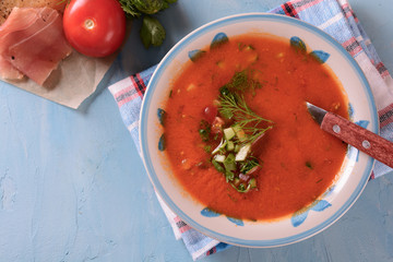Cold tomato soup gazpacho in a bowl next to sliced ​​jamon and vegetable ingredient on a light blue background, top view