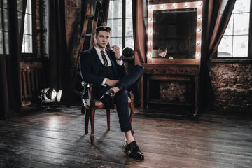 Fashion luxury man sitting on chair. Stylish businessman in full suit posing in interior room. Young handsome male model. Business concept. Success lifestyle