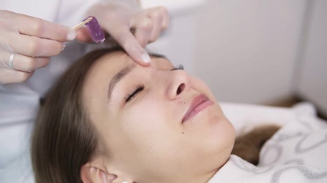 Girl lying on couch during eyebrowes treatment at studio beauty, beautician depilating and shaping brows at beauty salon, removing the wax between the brows