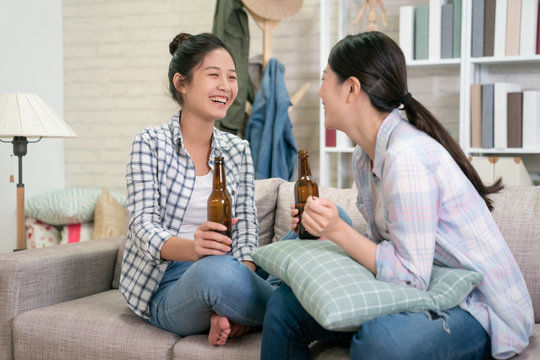 Two happy young female friends with beer bottles enjoying conversation in living room at home. girls roommate chatting on sofa in dormitory on weekends. asian women relax talk on couch in cozy house.