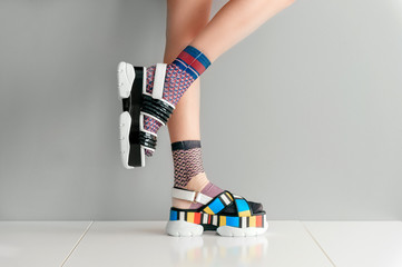 Beautiful female legs in mismatched trendy socks standing in two different fashionable high wedge leather sandals on white surface. Odd disargonized  young girl wearing high sole summer stylish shoes.