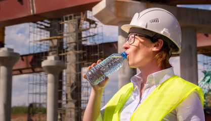 girl foreman drinks water from plastic bottle on background construction car road. man quenches...