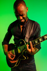 Young african-american musician playing the guitar like a rockstar on gradient green-yellow background. Concept of music, hobby, festival, open-air. Joyful attractive guy improvising, singing song.