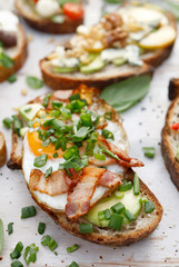 Open sandwich made of slices of sourdough bread with addition of avocado, egg, bacon, fresh scallion on a wooden white table, close-up. 