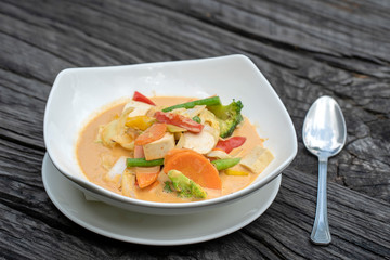 Mixed vegetables in creamy red curry coconut sauce with tofu in a white plate on a wooden table , traditional Thai food.