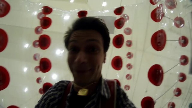 Attractive dark-haired guy with mix of facial emotions covered by soccer inflatable bubble is being dynamically rolled on plastic grass in small football field under white dome in amusement park.