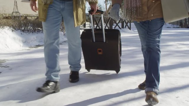 Close-up legs shot of man and woman, wearing jeans, boots and warm jackets, walking along snow-covered countryside hotel grounds towards entrance, wheeling suitcase, then going up stony steps
