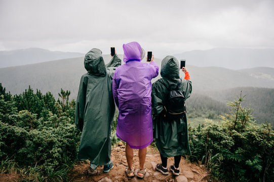 Group of travelers in raincoats stand high in the mountains and take photos of wild moody nature landscape with phones. Three wet adventure tourists make seilfie with smartphone in rainy summer day.