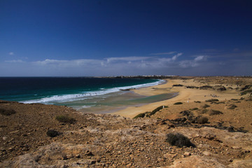 View over sand dunes with green ocean on (Playa del Aljibe) on white village on steep cliff (El Cotillo) - North Fuerteventura