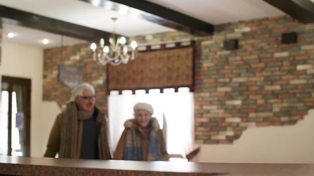 POV Shot Of Hotel Receptionist Standing Behind Front Desk, And Senior Caucasian Couple, Dressed In Winter Jackets, Walking Towards Her From Entrance, Greeting Her With Smiles And Giving Passports