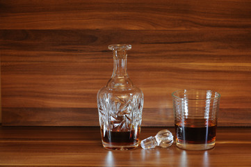 An open whiskey decanter, a glass of whiskey under a lamp on a wooden table.