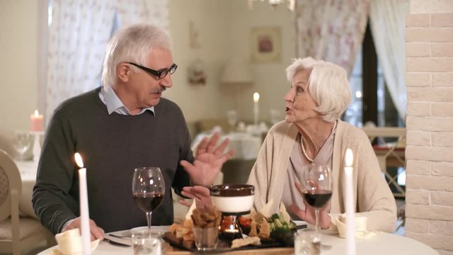 Waist-up shot of mature Caucasian couple dining together in hotel restaurant, with candles, snack platter and red wine, and talking animatedly to each other with hand gestures